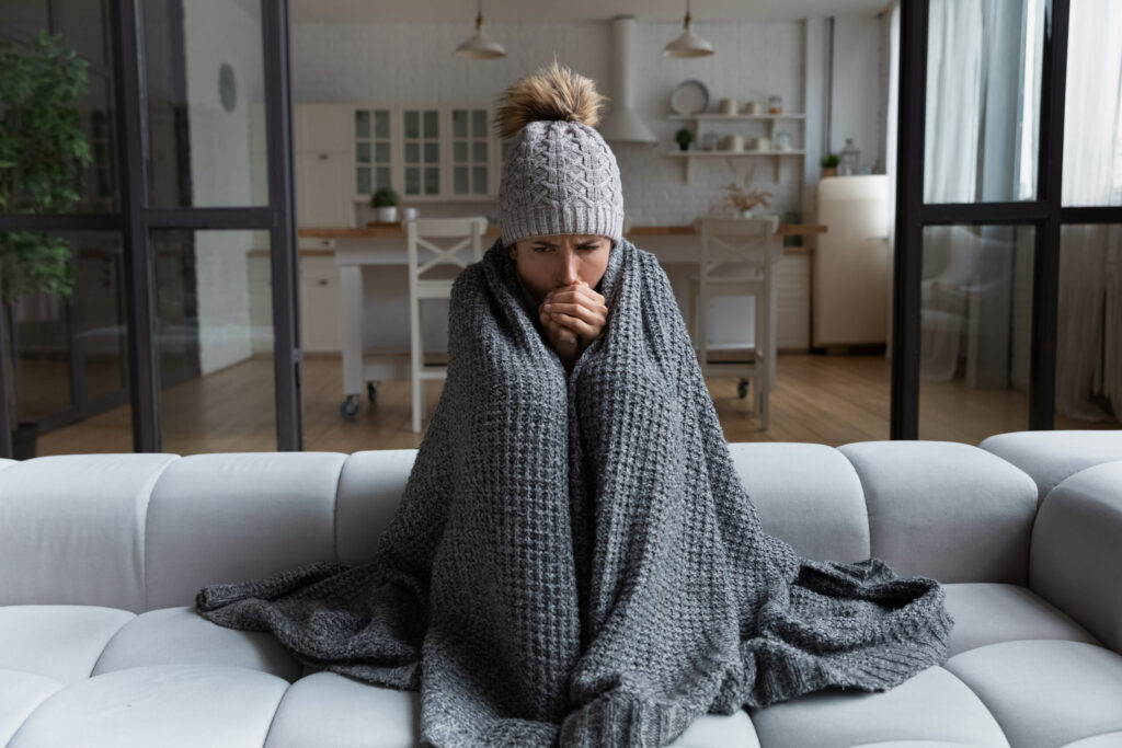 Woman sitting on a couch and bundled up in blankets due to a cold house from a malfunctioning thermostat